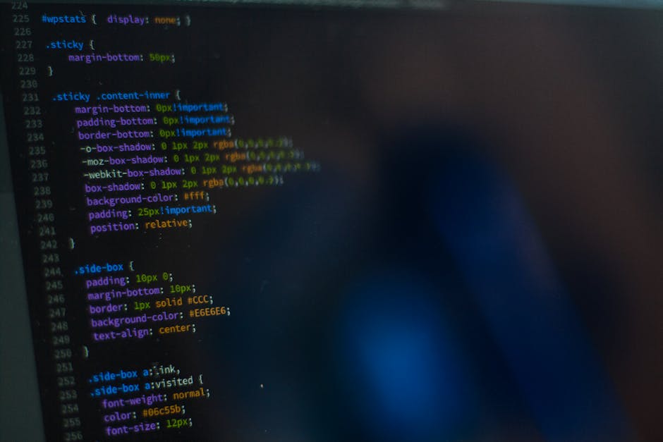A person working on a laptop with a dark screen displaying lines of computer code, situated on a black table indoors, possibly in an office or home environment. The individual’s hands are visible, w