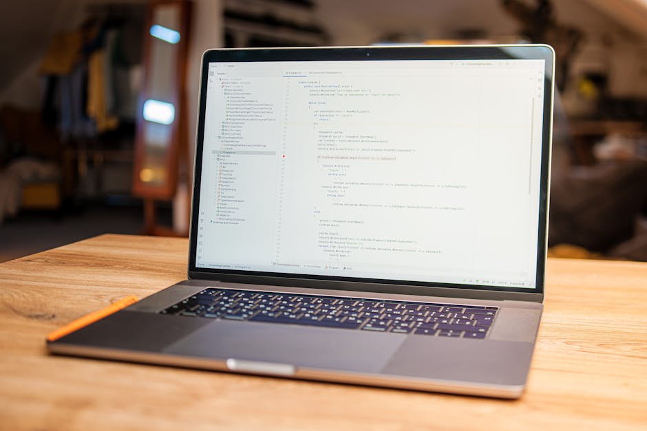 Close-up view of a laptop computer with a black keyboard and a red backlight, displaying a screen filled with lines of code in various colors, positioned on a desk or table. To the right of the laptop