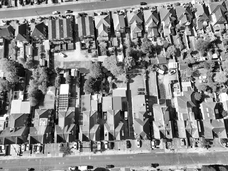 An aerial black and white photograph of a residential neighbourhood showing a row of terraced houses with pitched roofs, gardens, and driveways. The scene includes several parked cars along the street
