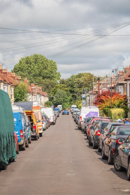 An aerial black and white photograph of a residential neighbourhood showing a row of terraced houses with pitched roofs, gardens, and driveways. The scene includes several parked cars along the street